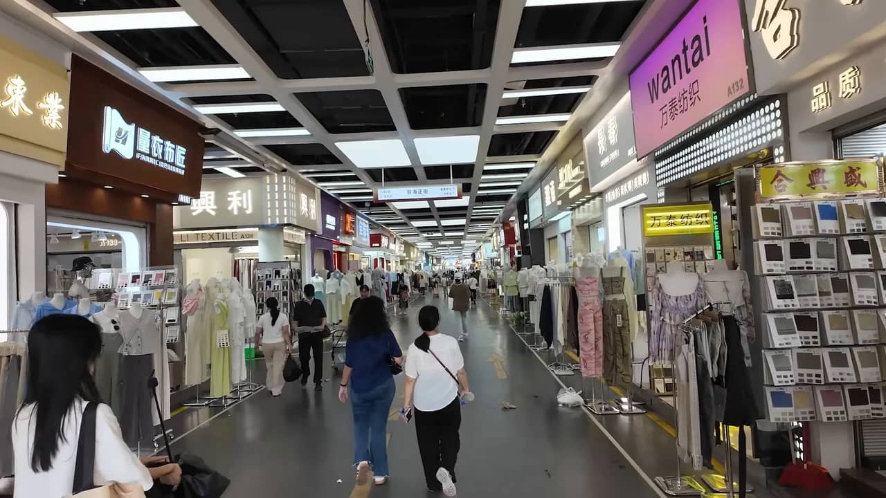 Allée d'un marché textile chinois, avec des présentoirs de tissus et des vêtements de chaque côté.