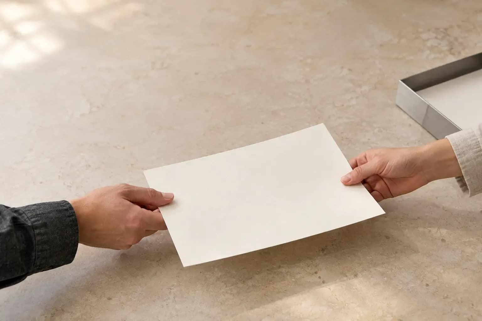 Deux mains se transmettent une feuille vierge au-dessus d'une table claire, près d'un plateau en aluminium.