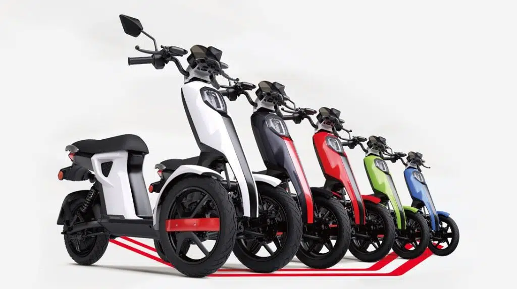 Quatre scooters électriques de couleurs différentes sont alignés sur un fond blanc. Les scooters sont fabriqués par différents fabricants et comportent trois roues pour une stabilité accrue.