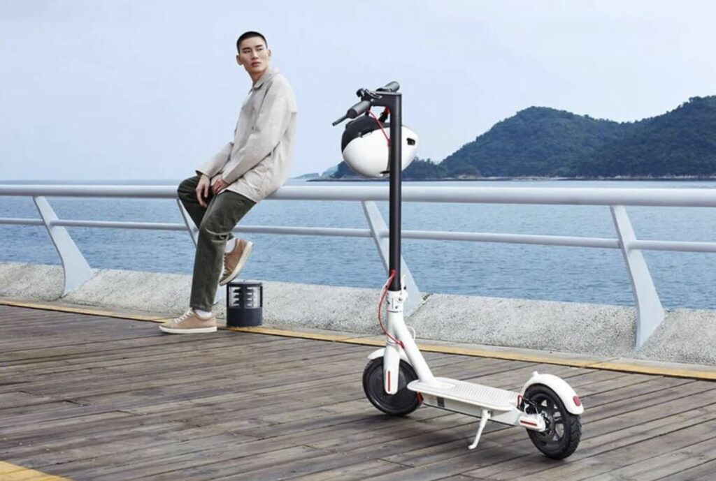 Un homme est assis sur un quai au bord de l’océan, avec un scooter électrique à ses côtés.